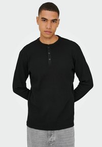 Sort langærmet henley skjorte med ribbet tekstur, rund hals og tre-knaps placket. Båret med grå jeans, hænderne placeret bag ryggen.