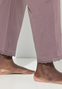 Volné levandulové kalhoty se zubatým krajkovým lemováním na kotnících, vyrobené z měkké tkaniny, s bosýma nohama na světlém povrchu.