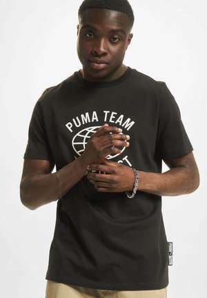 Svart bomull t-skjorte med hvit "PUMA TEAM" logo i sirkulært design, korte ermer, og etikett på venstre side. Modellen står med hendene foldet.