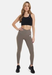 Zwarte sportbh en beige high-waisted leggings met een gladde textuur. Witte logo-accent op de legging. Zwarte hardloopschoenen.
