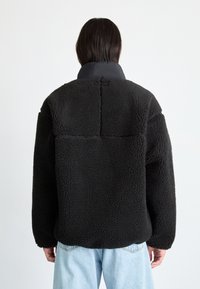 Schwarze Fleecejacke mit hohem Kragen, strukturiertem Stoff, lässiger Passform und sichtbaren Nähten, kombiniert mit hellblauen Jeans.