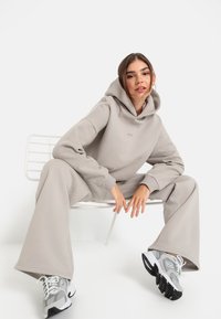 Svetlo šedá mikina s kapucňou a zodpovedajúce široké nohavice vyrobené z mäkkého materiálu. Model má na sebe šedé a biele športové topánky a sedí na bielej stoličke.