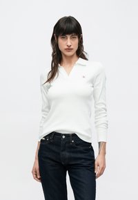 Biele polo tričko s dlhým rukávom v rebrovanej textúre s V-krkom a logom CK na ľavej strane hrude, v kombinácii s tmavomodrými rifľami.