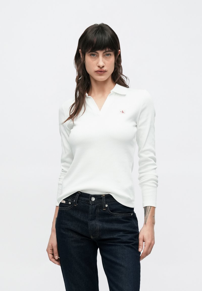 Biele polo tričko s dlhým rukávom v rebrovanej textúre s V-krkom a logom CK na ľavej strane hrude, v kombinácii s tmavomodrými rifľami.