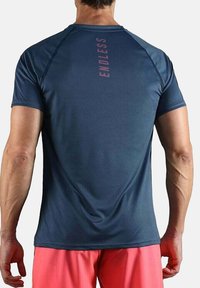 Hombre de espaldas con una camiseta atlética azul marino con el texto rojo "ENDLESS" en vertical en la parte superior de la espalda y pantalones cortos coral.