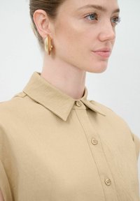 Camicia beige con colletto in tessuto cremoso, dotata di chiusura a bottoni e maniche corte. Orecchini ovali dorati come accessori di rilievo.