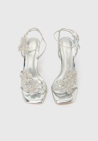 Ασημί σανδάλια με ψηλό τακούνι και τετράγωνη μύτη, διακοσμημένα με πολλαπλές ανθοειδείς λεπτομέρειες με στρας και λεπτά λουράκια στον αστράγαλο.