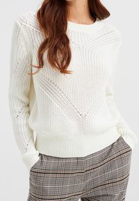 Vit stickad tröja med ett strukturerat mönster, rundad halsringning och ribbad nederkant. Matchad med bruna rutiga byxor i lös passform.