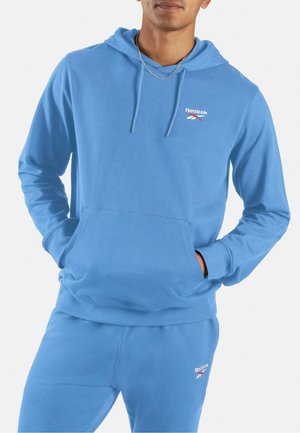Mann trägt blauen Reebok-Hoodie und passende Sweatpants, Hände in den Taschen des Hoodies, weißer Hintergrund.
