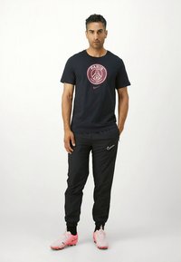 Donkerblauw T-shirt met een rode ronde Parijse logo, gecombineerd met zwarte sportbroek met een wit Nike-logo en roze voetbalschoenen.