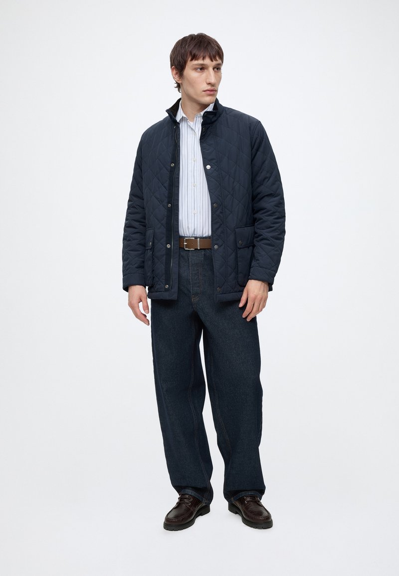 Mand iført en navy quiltet jakke, blåstribet skjorte, mørke løse jeans og brune sko står mod en ensfarvet lys baggrund.