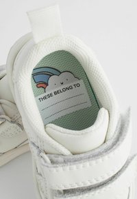 Witte kindersneakers met grijze accenten, voorzien van een gevoerde tong en een label binnenin met de tekst "DEZE HOREN BIJ" met een regenboogontwerp.