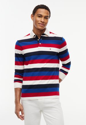 Jeune homme portant un polo à manches longues avec des rayures horizontales rouges, bleues, noires et blanches et un pantalon blanc, debout devant un fond blanc.