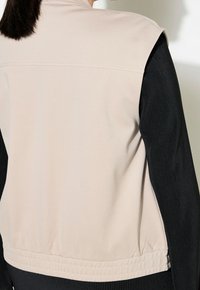 Beżowa, bezrękawnikowa kamizelka z elastycznym, czarnym, prążkowanym bokiem, wyposażona w subtelne przeszycia na górze i elastyczny dół dla dopasowanego kroju.