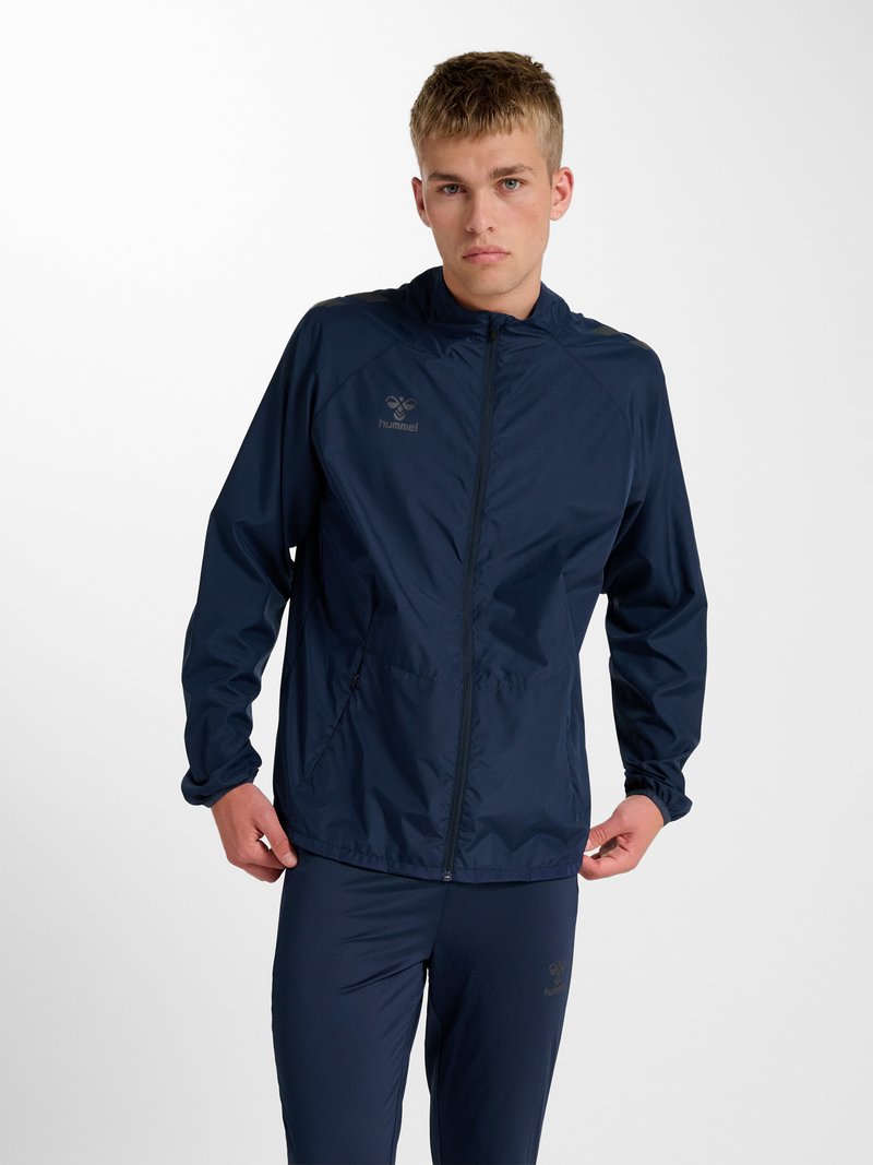 Giovane uomo con giacca sportiva blu navy con zip intera e pantaloni abbinati, che sistema l'orlo della giacca, in piedi davanti a uno sfondo bianco semplice.