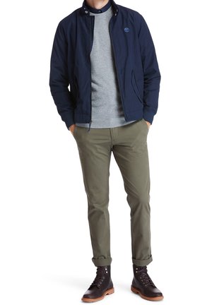 Mand iført marineblå jakke med lynlås, grå sweater, olivengrønne bukser med opkanten ved anklerne og mørkebrune snørestøvler, stående med hænderne i lommerne.