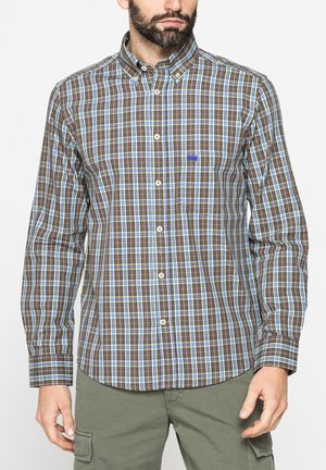 Uomo che indossa una camicia a quadri marrone, blu e bianca con taschino sul petto e pantaloni cargo verde oliva, in piedi su sfondo bianco.