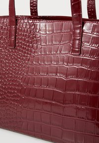 Sac à main bordeaux en matériau synthétique imitation crocodile brillant, avec une surface texturée présentant des écailles petites et grandes, et deux poignées robustes.