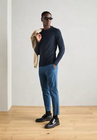 Navystrukturierter Strickpullover, blaue schmal geschnittene Hose, schwarze Schnürschuhe und Sonnenbrille. Helle beige Jacke über die Schulter gelegt.
