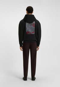 Uomo che indossa un hoodie nero con un grafico astratto rosso e grigio sul retro, pantaloni marrone scuro e scarpe nere, in piedi di spalle su uno sfondo grigio.