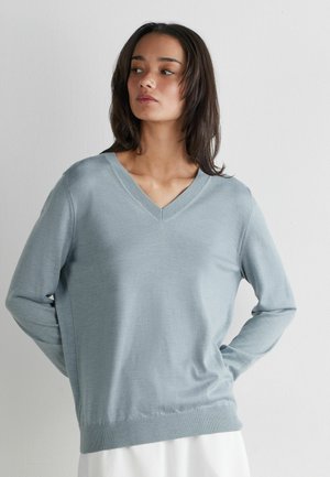 Kobieta o ciemnych włosach, ubrana w jasnoniebieski sweter z dekoltem w serek oraz białą spódnicę, stoi na tle gładkiej, jasnej ściany.