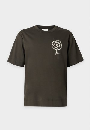Ung mand iført sort t-shirt med hvidt spiraldesign, lyseblå vide jeans og mørkebrune sko, stående med den ene hånd i lommen.