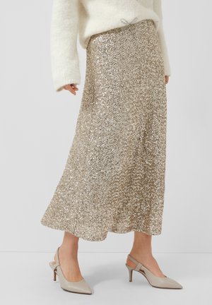 Frau in einem schimmernden, champagnerfarbenen Pailletten-Midirock, weißem, flauschigem Pullover und beigen Slingback-Kitten-Heels, steht vor einem weißen Hintergrund.