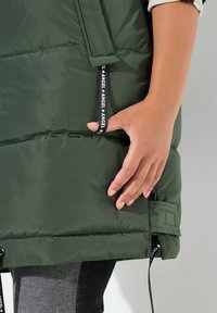 Gilet imbottito verde scuro con colletto alto e chiusura con zip. Presenta tasche laterali con tiranti con logo e un design trapuntato per l'isolamento.