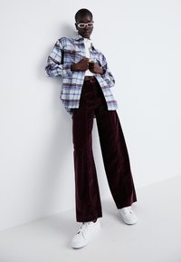 Cămașă flanel cu carouri în nuanțe de albastru și alb, butonată, asortată cu pantaloni largi din corduroy în nuanța burgundă și adidași albi. Accesoriile includ ochelari de soare albi.