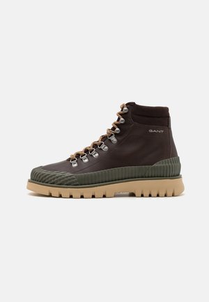 Brown leather high-top boots with green rubber accents, textured toe cap, metal eyelets, and tan laces, featuring a chunky outsole.