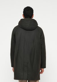 Homem de costas, vestido com um longo casaco preto com capuz e uma fenda central nas costas, em pé contra um fundo branco liso.