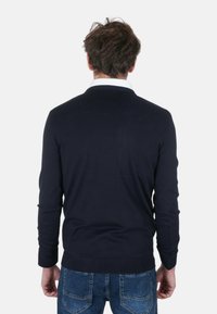 Maglione blu navy con scollo rotondo e polsini a coste, tessuto strutturato, indossato sopra una camicia bianca con colletto, abbinato a jeans di denim blu.