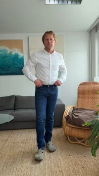 Cămașă albă cu guler, blugi albaștri și saboți gri. Stând într-un living cu o canapea gri, un covor bej deschis și o plantă în ghiveci.