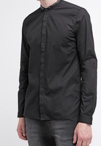 Camicia nera a maniche lunghe con colletto alla mandarino, texture liscia e chiusura a button, caratterizzata da una leggera lucentezza. Portata con pantaloni grigio scuro.