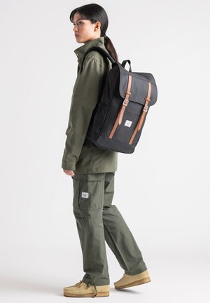 Black backpack with brown leather straps, rectangular shape, adjustable buckles, and a front label. Model wears an olive green outfit.