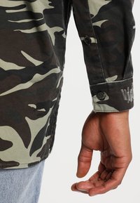 Camicia con motivo mimetico verde e nero, caratterizzata da polsini con bottoni e un dettaglio a bordo grezzo. Il tessuto sembra resistente con una texture morbida.