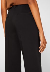 Femme portant un pantalon noir taille haute et un haut marron rentré, vue de dos avec la main droite reposant sur le côté.