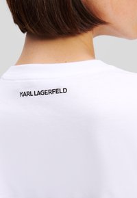 Bílý bavlněný tričko s kulatým výstřihem, na zadní části krku s černým potiskem loga "KARL LAGERFELD". Hladká textura, casual design.