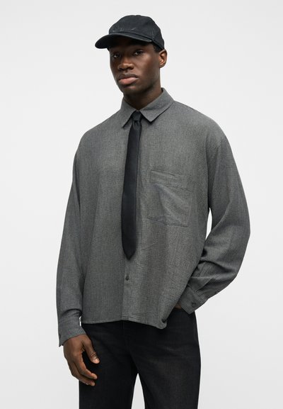 Άνδρας με μαύρο καπέλο, γκρι καρό πουκάμισο με τσεπάκι στο στήθος, μαύρη γραβάτα και μαύρο παντελόνι, στέκεται μπροστά σε απλό λευκό φόντο.