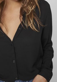 Blouse noire avec un col en V et des manches longues, dotée d'une fermeture à boutons sur le devant et d'un tissu lisse et texturé. Agrémentée de boutons ronds.