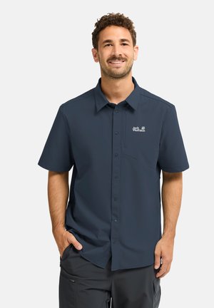 Uomo che indossa una camicia blu navy a maniche corte con bottoni e logo Jack Wolfskin, in piedi con una mano in tasca, sorridente su sfondo bianco.