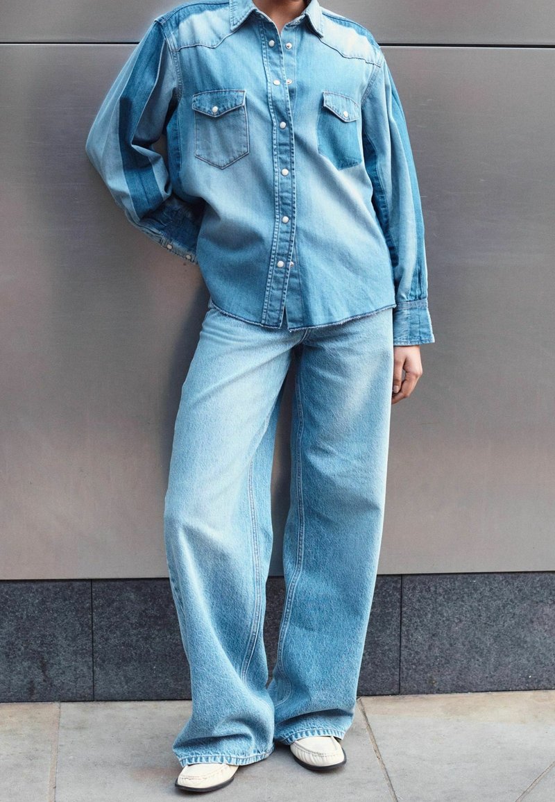 Personne portant une chemise en denim bleu clair et un jean à jambes larges, debout contre un mur gris, chaussures blanches visibles en bas.