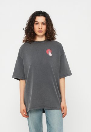 Άτομο με κυματιστά μαλλιά που φοράει μεγάλη σκούρα γκρι μπλούζα με μικρό ροζ σχέδιο γάτας στο στήθος, συνδυασμένη με ανοιχτό μπλε τζιν.