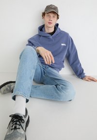 Pullover hoodie in blue cotton with a front pocket, paired with light blue denim jeans, and gray speckled shoes. Simple cap on the head.