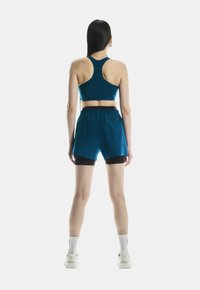 Teal sportbh met racerback ontwerp, gecombineerd met bijpassende teal shorts met een zwarte binnenlaag. Model draagt witte enkelsokken en sneakers.
