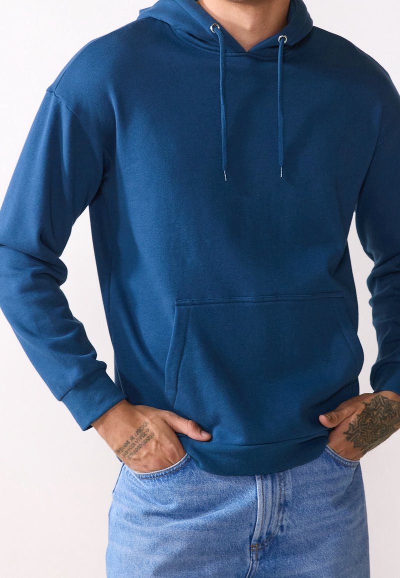 Homme portant un sweat à capuche bleu avec une poche avant et un jean bleu clair, mains partiellement dans les poches, tatouages visibles sur les avant-bras.