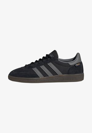 Baskets Adidas noires avec des bandes grises, des lacets noirs, des matériaux en suède et en mesh, et une semelle marron texturée.