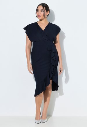 Femme en robe portefeuille bleu marine avec détails à volants et talons hauts argentés, debout devant un fond blanc uni.