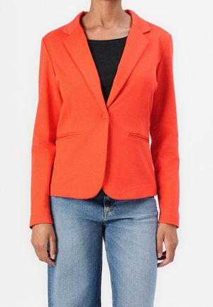Vrouw draagt een feloranje blazer over een zwarte top en blauwe spijkerbroek, staand tegen een effen witte achtergrond.