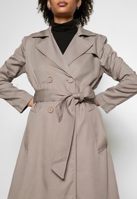 Trench coat doppiopetto beige con vita affusolata, colletto deciso e due tasche laterali. Tessuto liscio con una silhouette su misura.